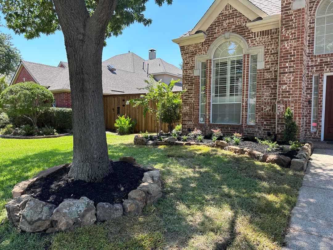 Brick house exterior with landscaped garden, tree, and rock border in a sunny neighborhood.