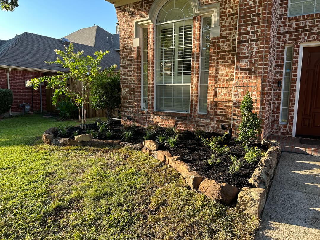 Lush landscaped garden with stone border, featuring green plants by a brick home entrance.