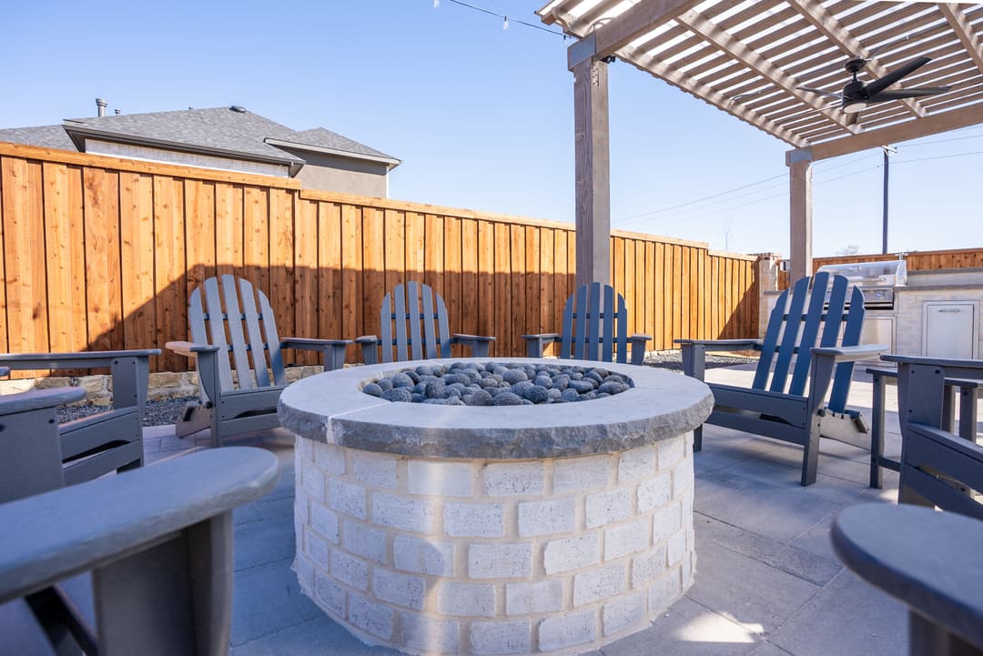 Outdoor fire pit with seating around it, surrounded by a wooden fence and blue sky.