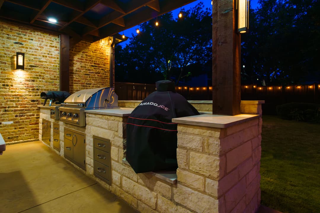 Outdoor kitchen with barbecue grill and Kamado Joe smoker, illuminated at night.