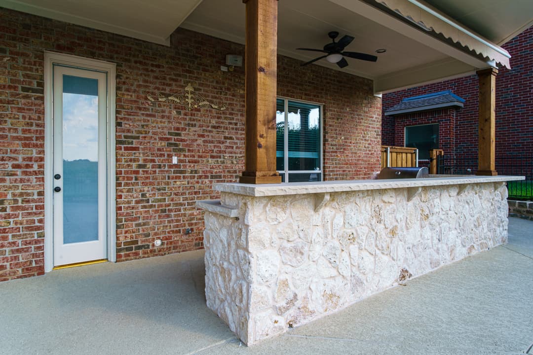 Outdoor patio with stone bar, wooden pillars, and a door leading to a brick house.
