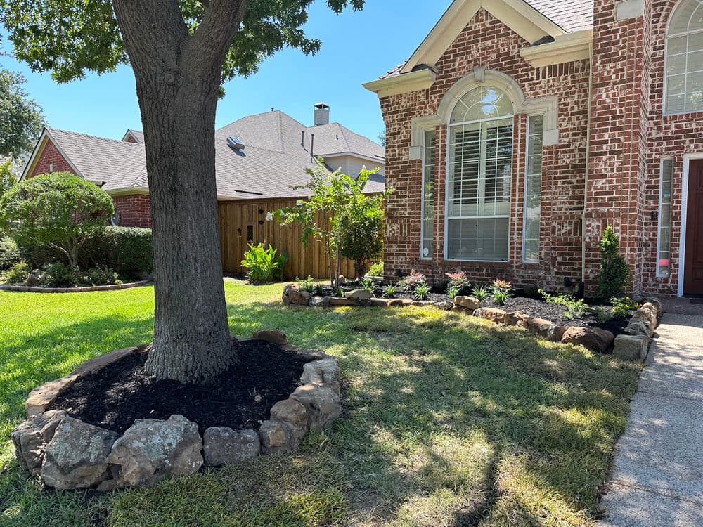 Front yard landscape featuring a brick house, tree, and vibrant garden with rocks and mulch.