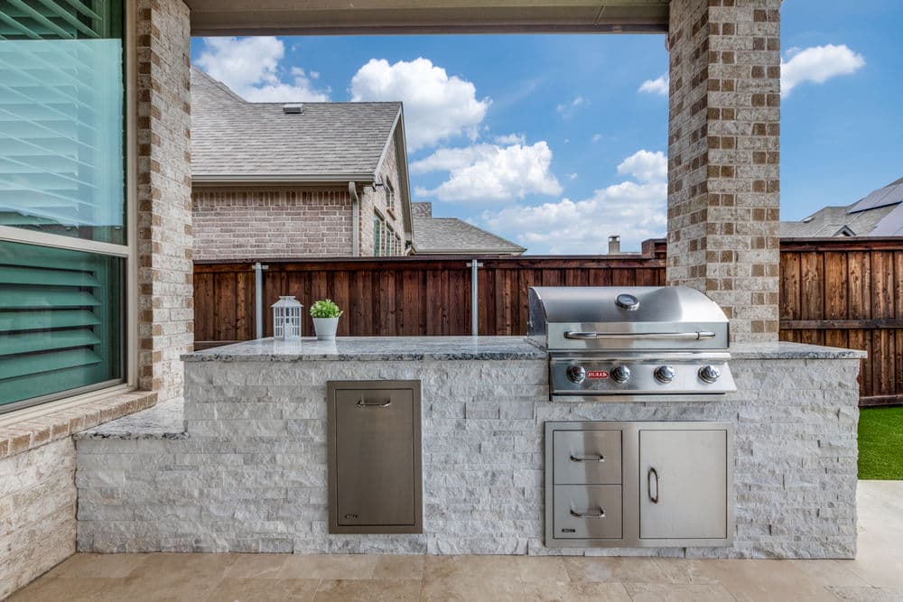 Modern outdoor kitchen with stainless steel grill and granite countertop in backyard setting.